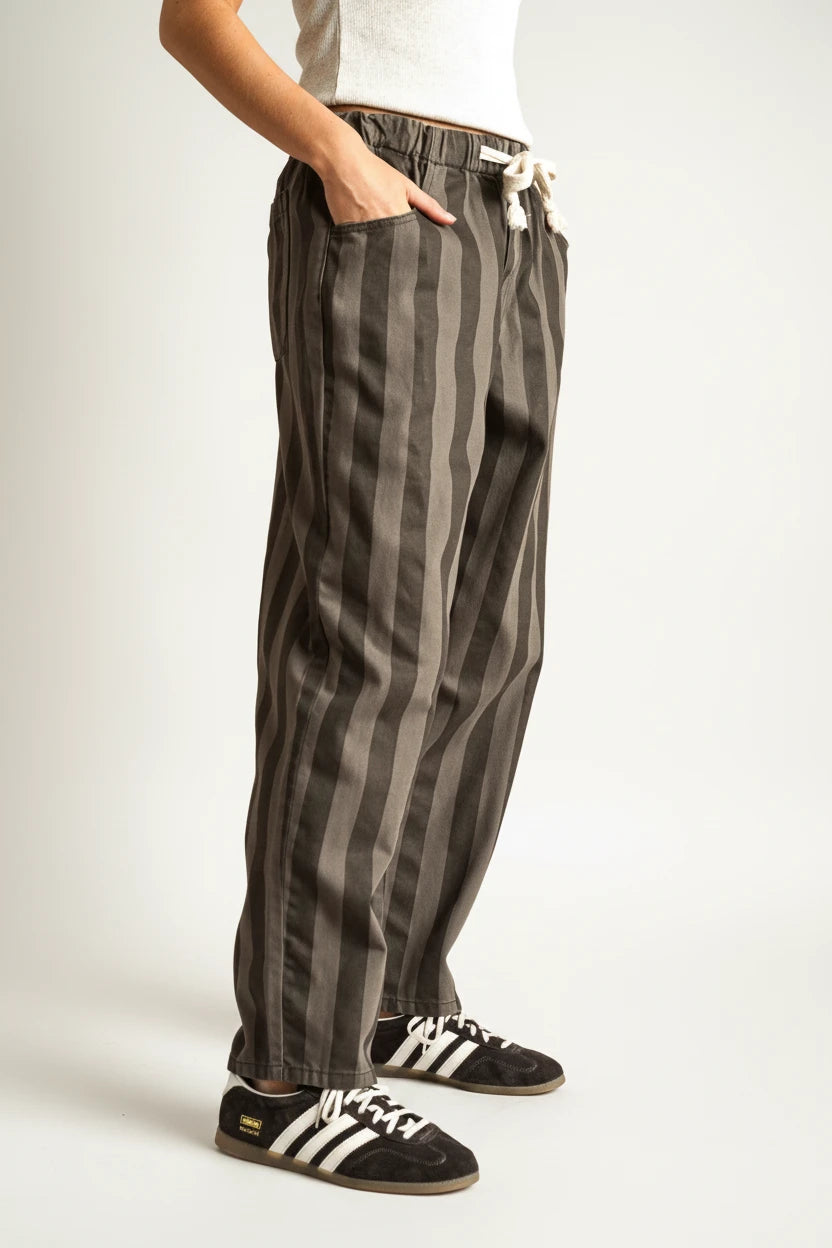 A person wearing loose vertical-striped charcoal gray pants with a white drawstring, paired with black sneakers, shown from the waist down against a solid beige studio background.