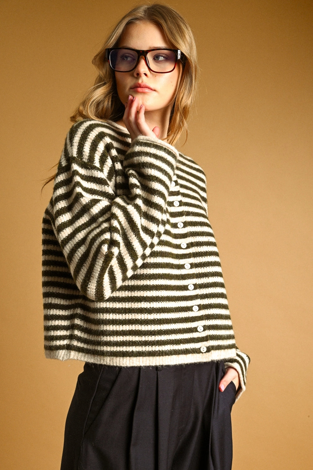 A woman wearing thick black-framed glasses and an oversized black and cream striped knit button-front top, posed against a warm tan studio background.