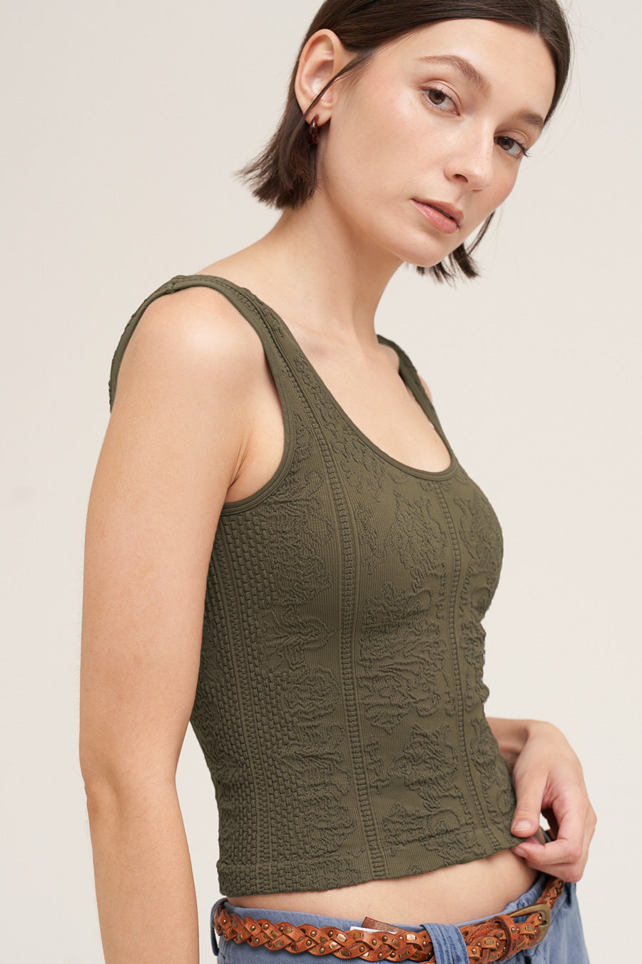 A woman wearing a textured olive green ribbed tank top, paired with blue jeans and a brown braided belt, photographed against a neutral light beige background.