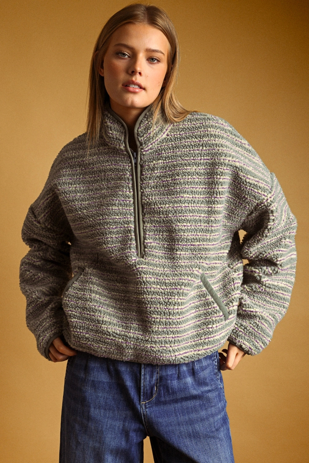 A woman wearing a textured, heathered gray and white quarter-zip fleece pullover sweater with blue jeans, posed against a solid warm brown studio background.