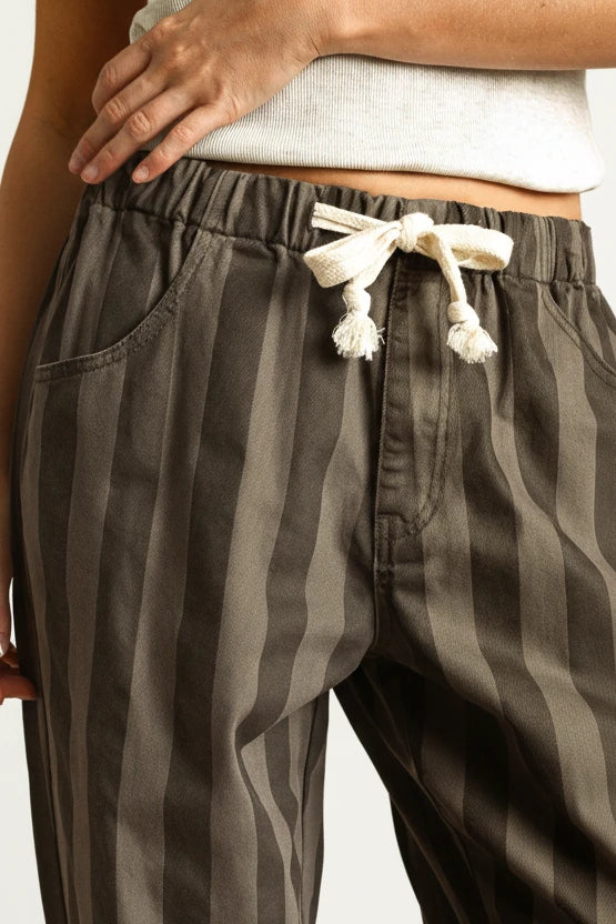 Close-up of a person wearing dark gray and brown vertically striped drawstring pants with a cream-colored drawstring, paired with a light gray ribbed top against a warm beige background.