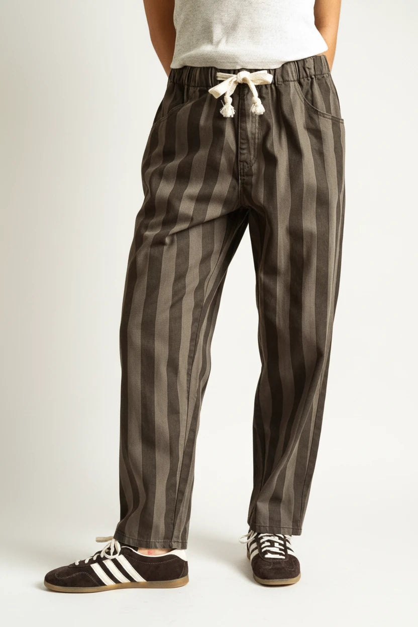 A person wearing loose vertical-striped drawstring pants in dark gray and black, paired with a white top and black sneakers, against a warm tan studio background.
