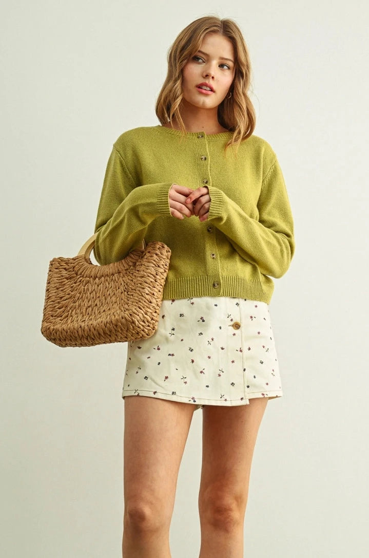 Woman wearing a green sweater and white skirt with a straw bag against a light background