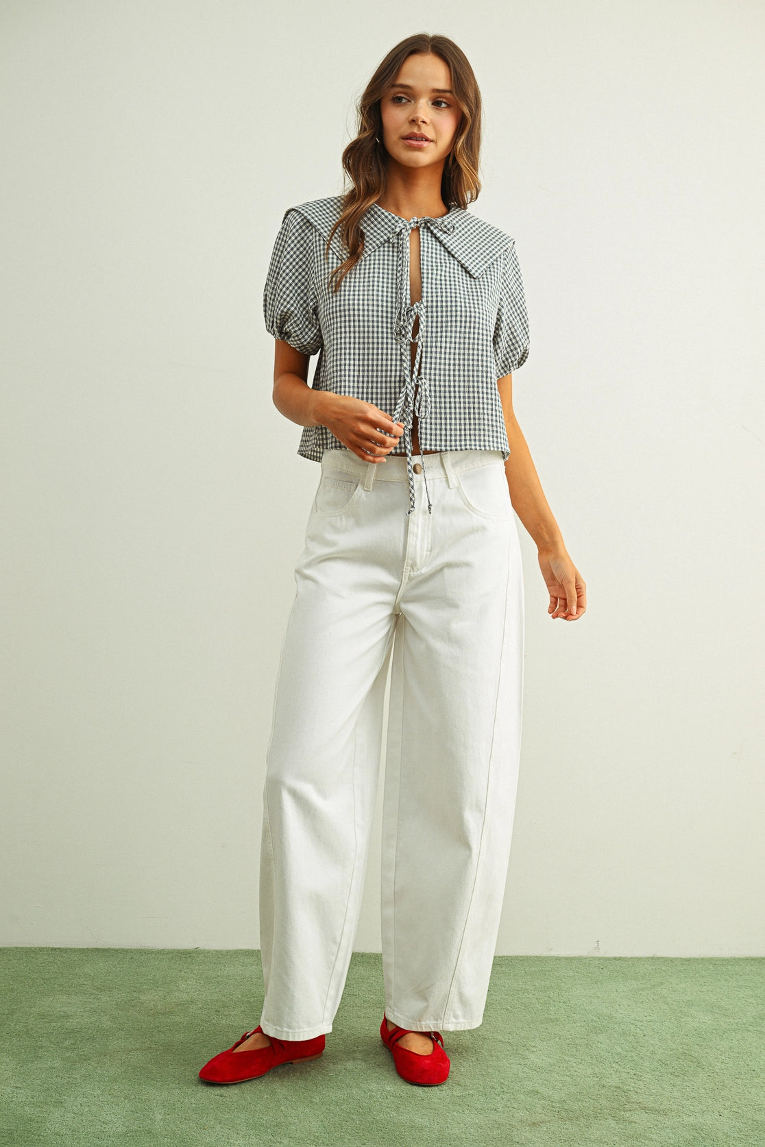 Woman wearing a checkered blouse, white pants, and red shoes on a plain background