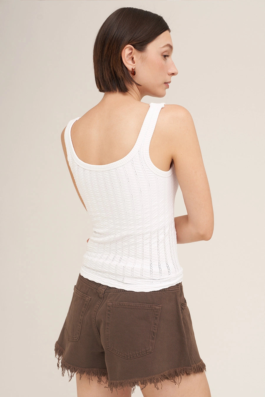 A woman shown from the back wearing a fitted white ribbed tank top and brown shorts against a plain light beige studio background.