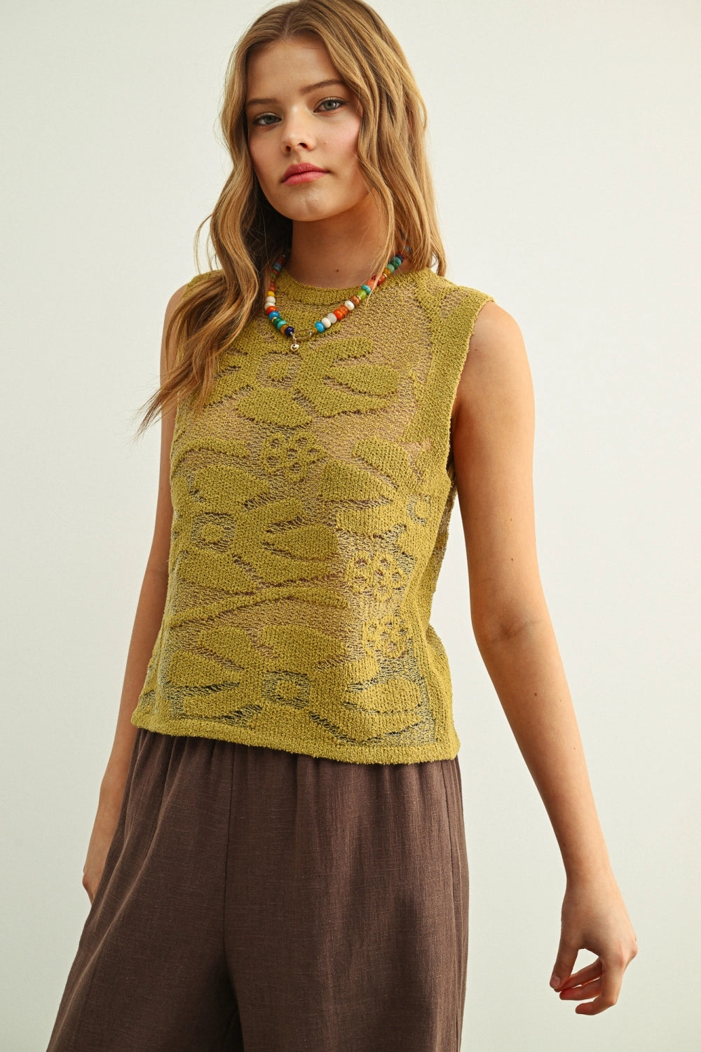 Woman wearing a yellow sleeveless top and brown pants against a plain background