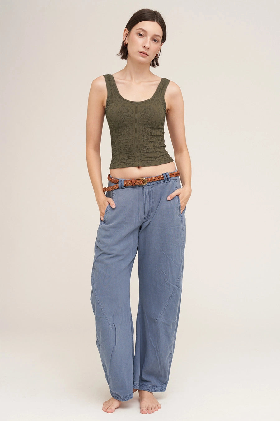 A model stands against a light beige studio background wearing a dark olive green ribbed tank top and wide-leg blue jeans with a brown patterned belt.