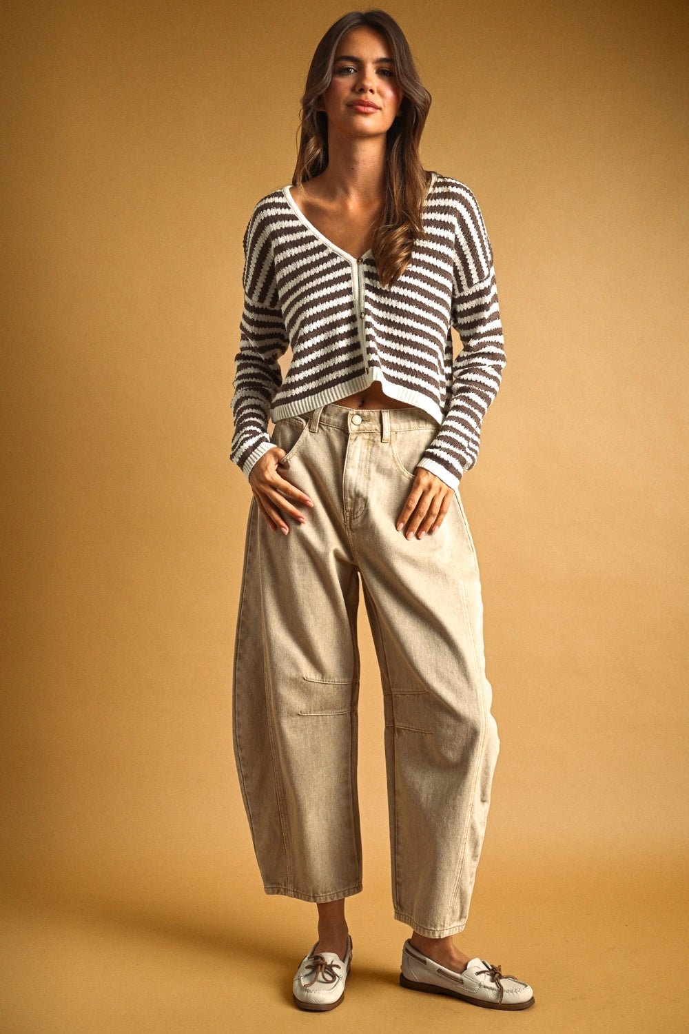 A woman stands against a warm tan studio backdrop wearing a cropped black-and-white striped knit cardigan, loose beige wide-leg pants, and white loafers.