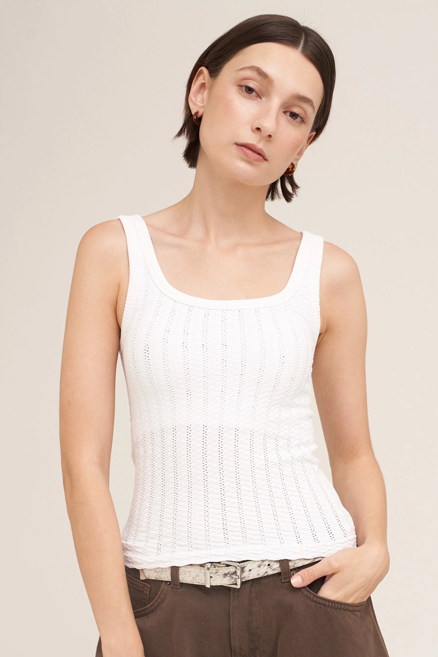 A woman wearing a white ribbed knit tank top with a scoop neckline, paired with brown pants and a patterned belt, against a solid light beige background.