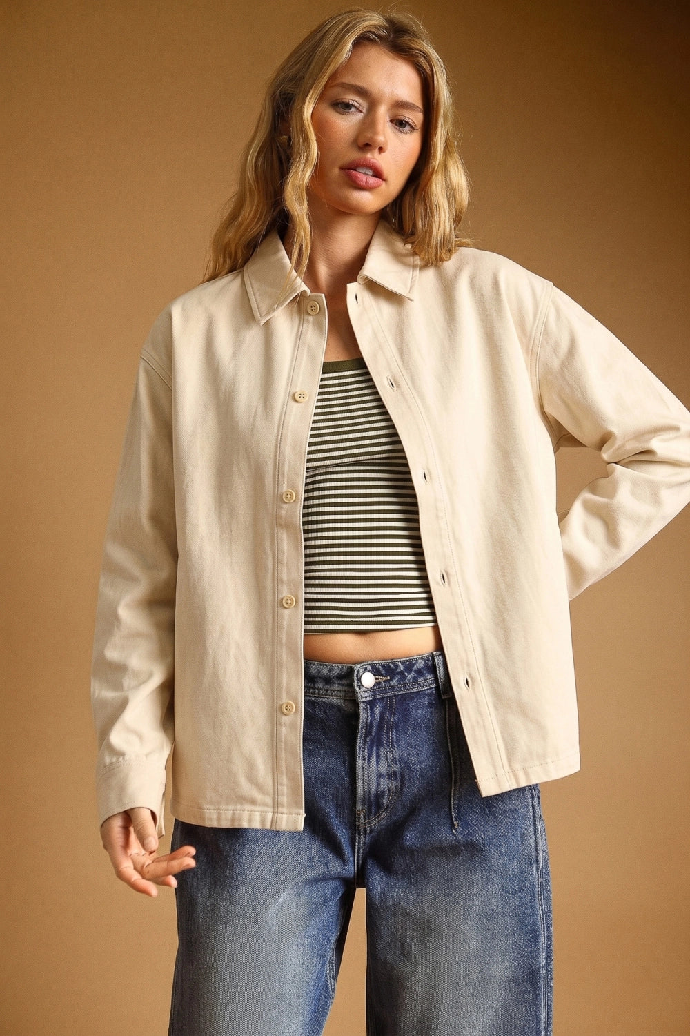 A woman modeling a light beige button-up overshirt over a black and white striped crop top and blue jeans, set against a solid warm brown studio background.