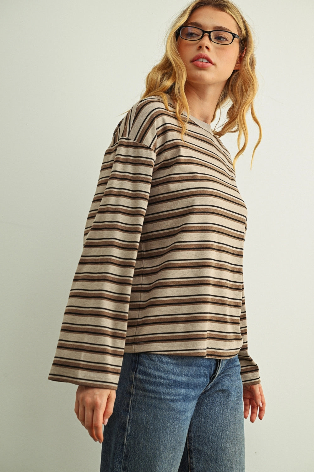 A woman wearing a beige and brown horizontally striped long-sleeve top with blue jeans, photographed against a plain light gray studio background.