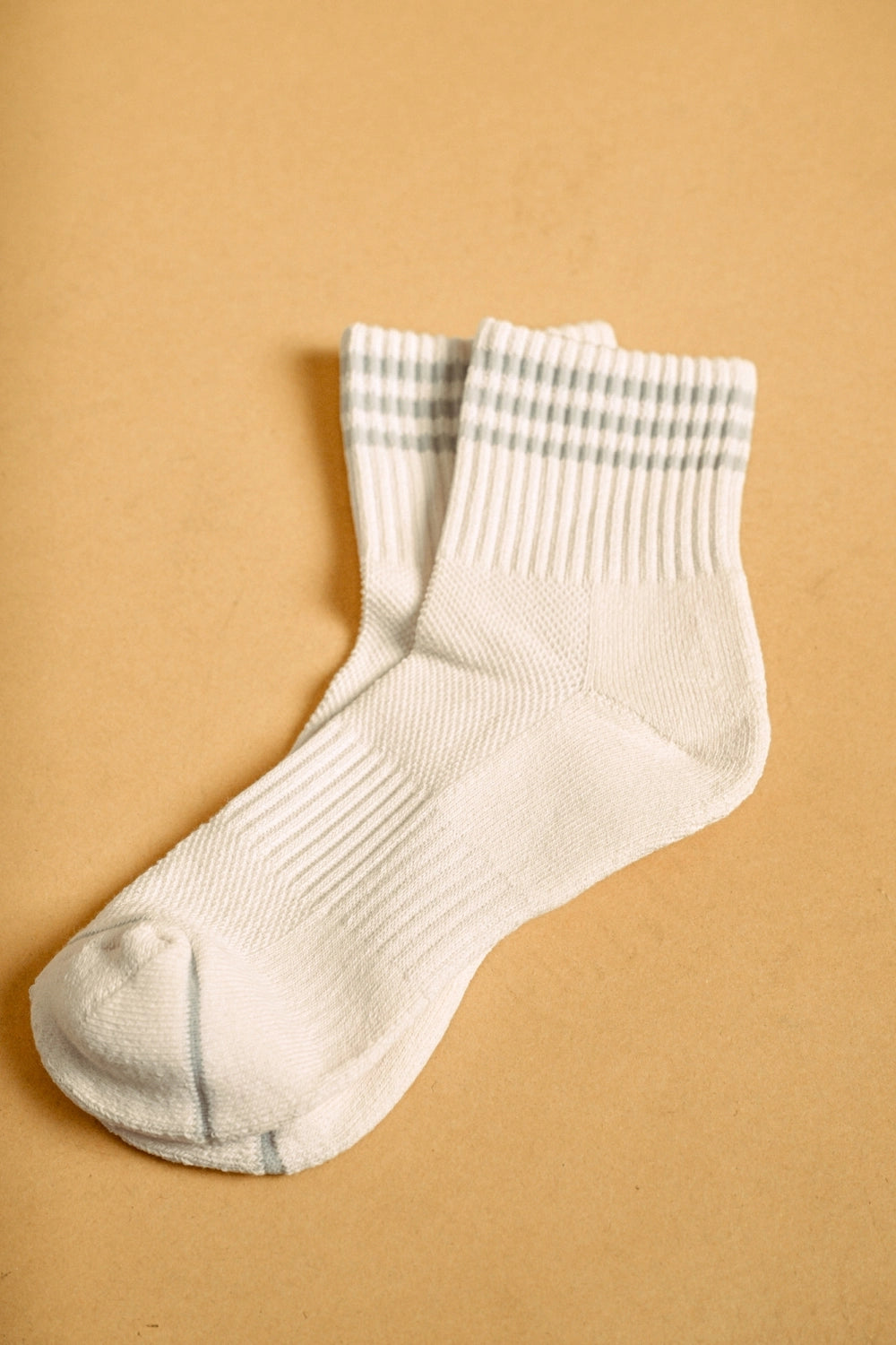 A pair of off-white crew socks with thin gray stripes at the ribbed cuffs, displayed on a warm tan background.