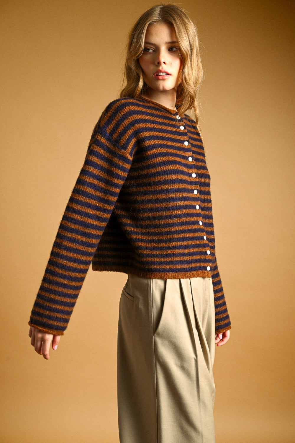 A woman modeling a cropped, long-sleeve knit cardigan with horizontal dark brown and rust-orange stripes, featuring a button-up front, against a solid warm tan studio background.