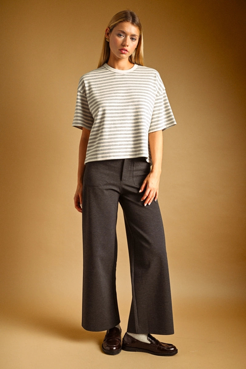 A woman stands against a warm tan studio background wearing a cropped short-sleeve striped top in off-white and gray, dark charcoal wide-leg pants, and dark brown loafers.