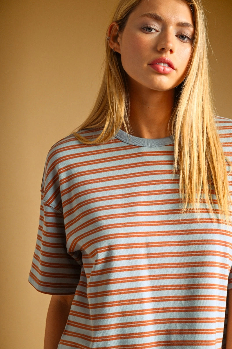 A woman wearing a short-sleeve crewneck t-shirt with thin horizontal rust-orange and light blue stripes, set against a solid warm beige background.
