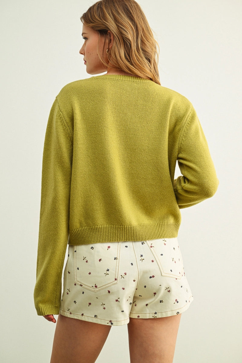 Woman wearing a green sweater and white shorts with a floral pattern on a plain background