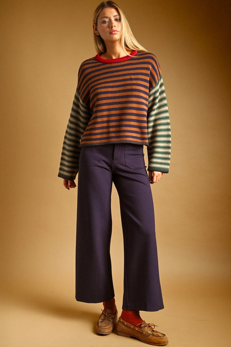 A woman modeling a striped knit sweater with green and orange horizontal stripes and solid green sleeves, paired with dark wide-leg trousers, red socks, and brown loafers against a warm brown studio background.