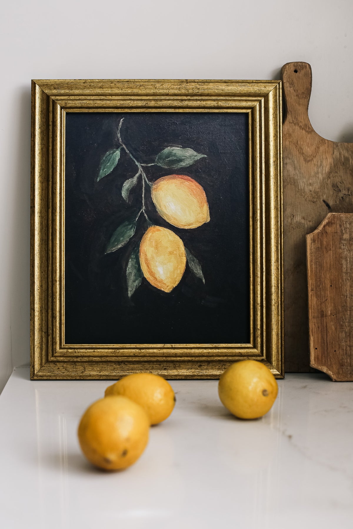 A framed art print of lemons on a dark background displayed on a table with lemons in front and a cutting board to the side.