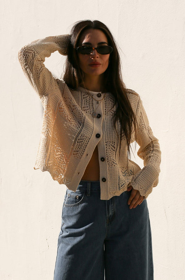 Woman wearing a beige knitted cardigan and sunglasses against a plain background
