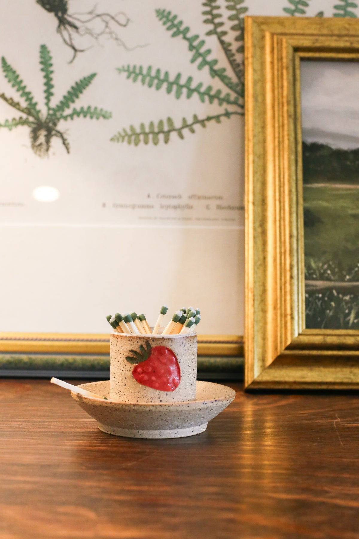 A beige ceramic strawberry match striker with a red strawberry design on the side, containing matches, placed on a wooden surface.