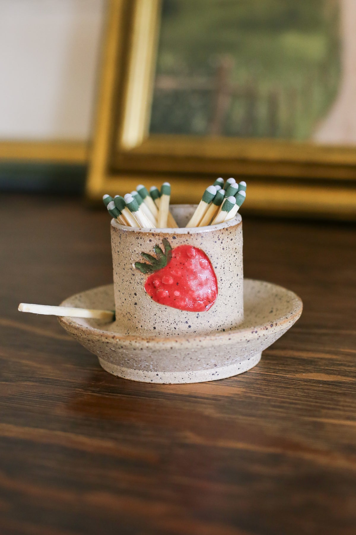 A beige ceramic strawberry match striker with a red strawberry design on the side, containing matches, placed on a wooden surface.
