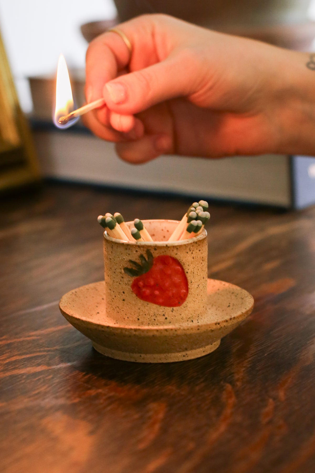 A beige ceramic strawberry match striker with a red strawberry design on the side, containing matches, placed on a wooden surface.