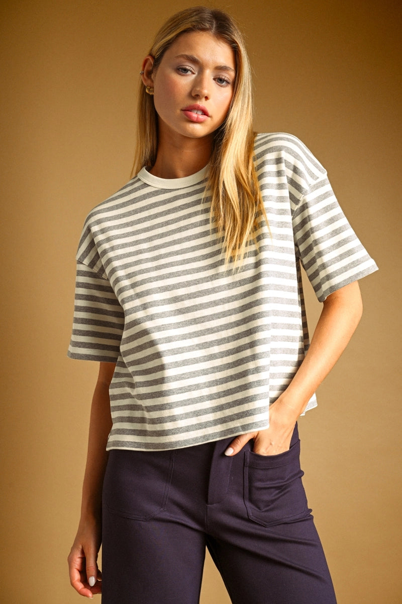 A woman modeling a short-sleeve crewneck t-shirt with horizontal gray and white stripes, paired with dark purple pants, set against a solid warm brown studio background.