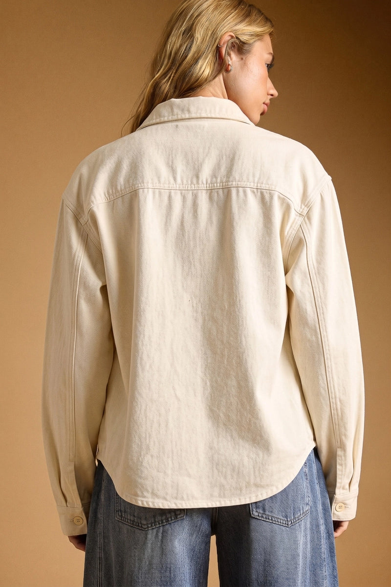 A woman shown from the back wearing a light cream-colored linen button-up shirt and blue jeans, set against a solid warm brown studio background.
