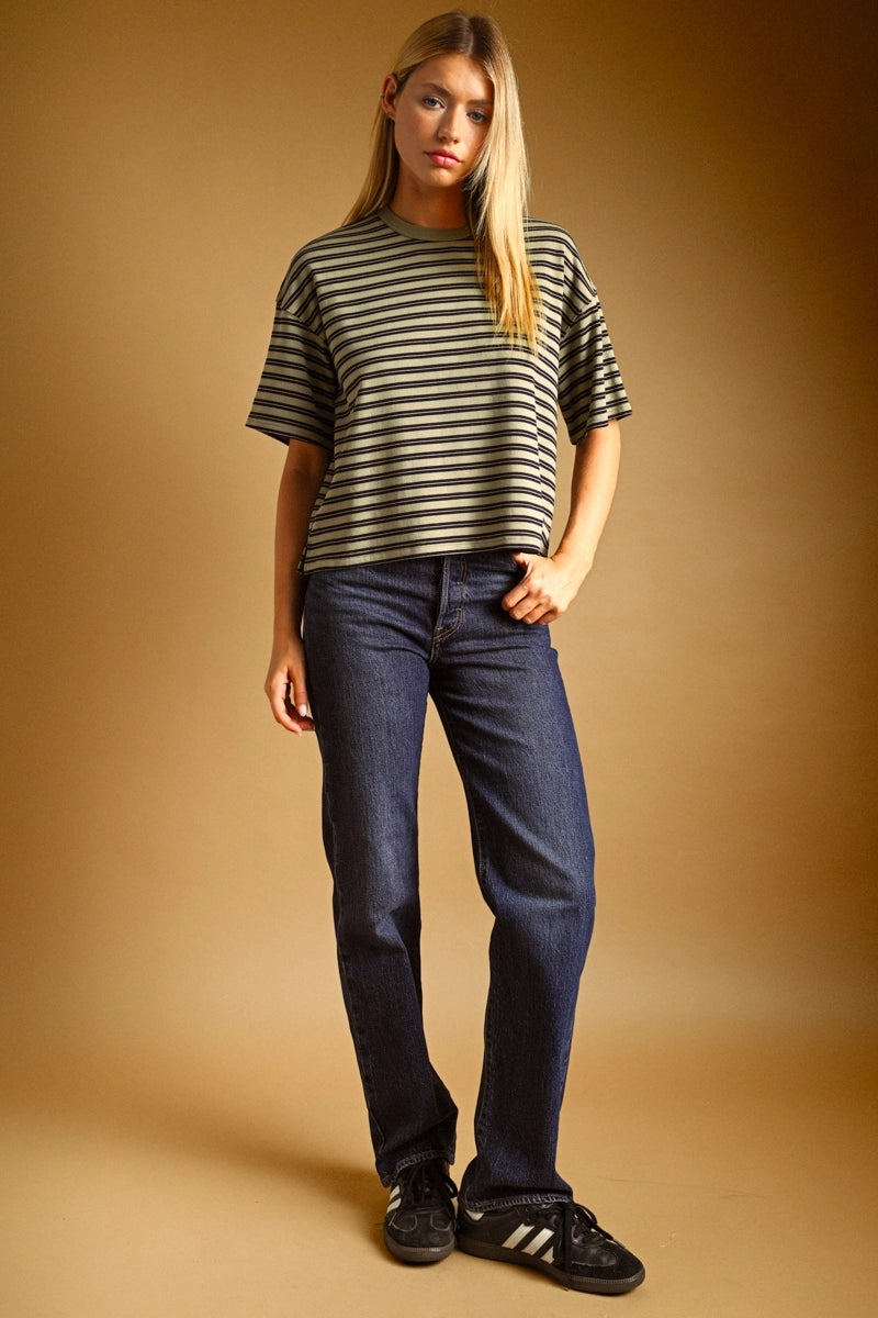 A woman modeling a short-sleeve striped t-shirt in olive green and black, paired with dark blue jeans and black sneakers, against a solid warm brown studio backdrop.