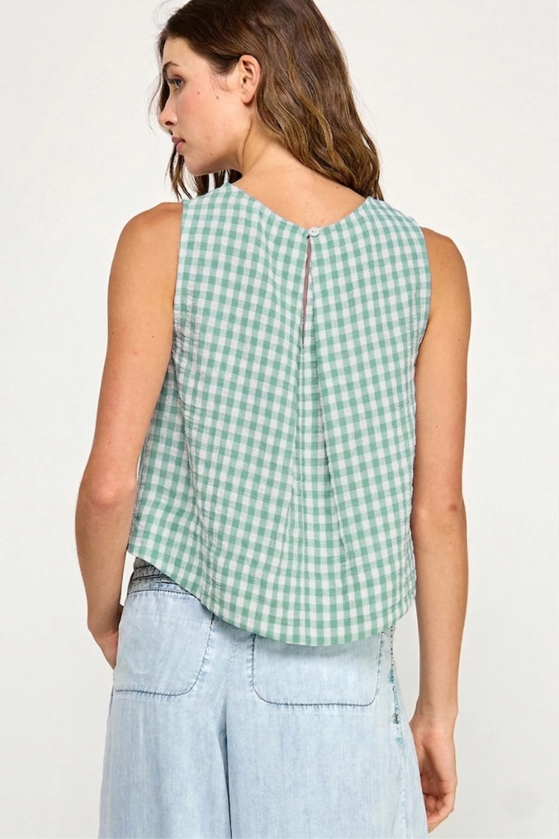 Woman wearing a green and white checkered sleeveless top with light blue pants on a plain background