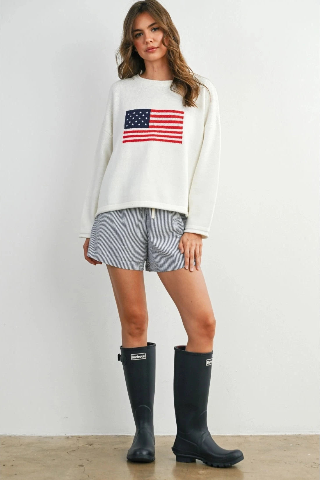 Woman wearing a white sweatshirt with an American flag design, plaid shorts, and black rain boots on a plain background.