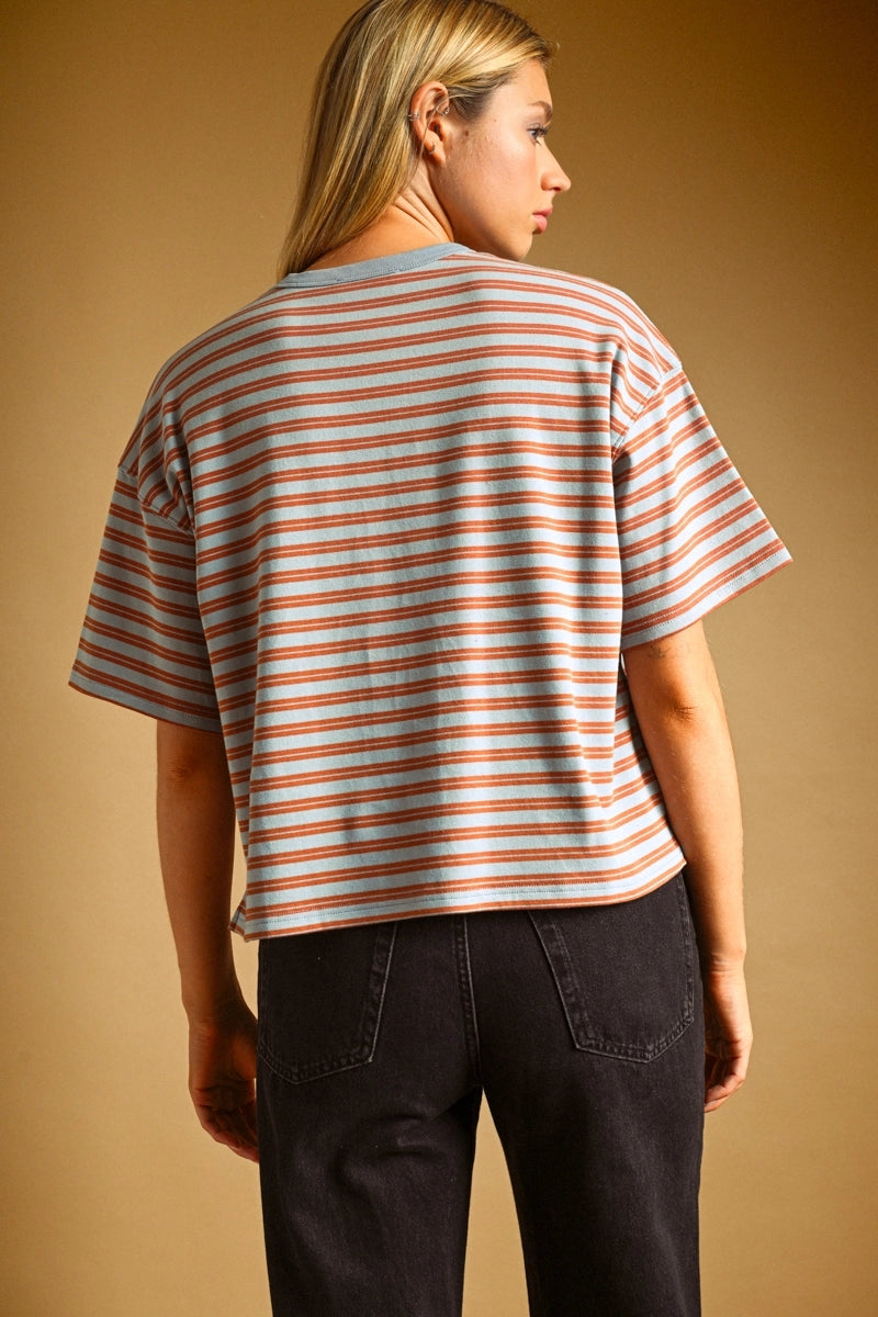 A woman shown from the back wearing a short-sleeve striped t-shirt with horizontal peach and gray stripes, paired with dark jeans, against a solid warm brown studio background.