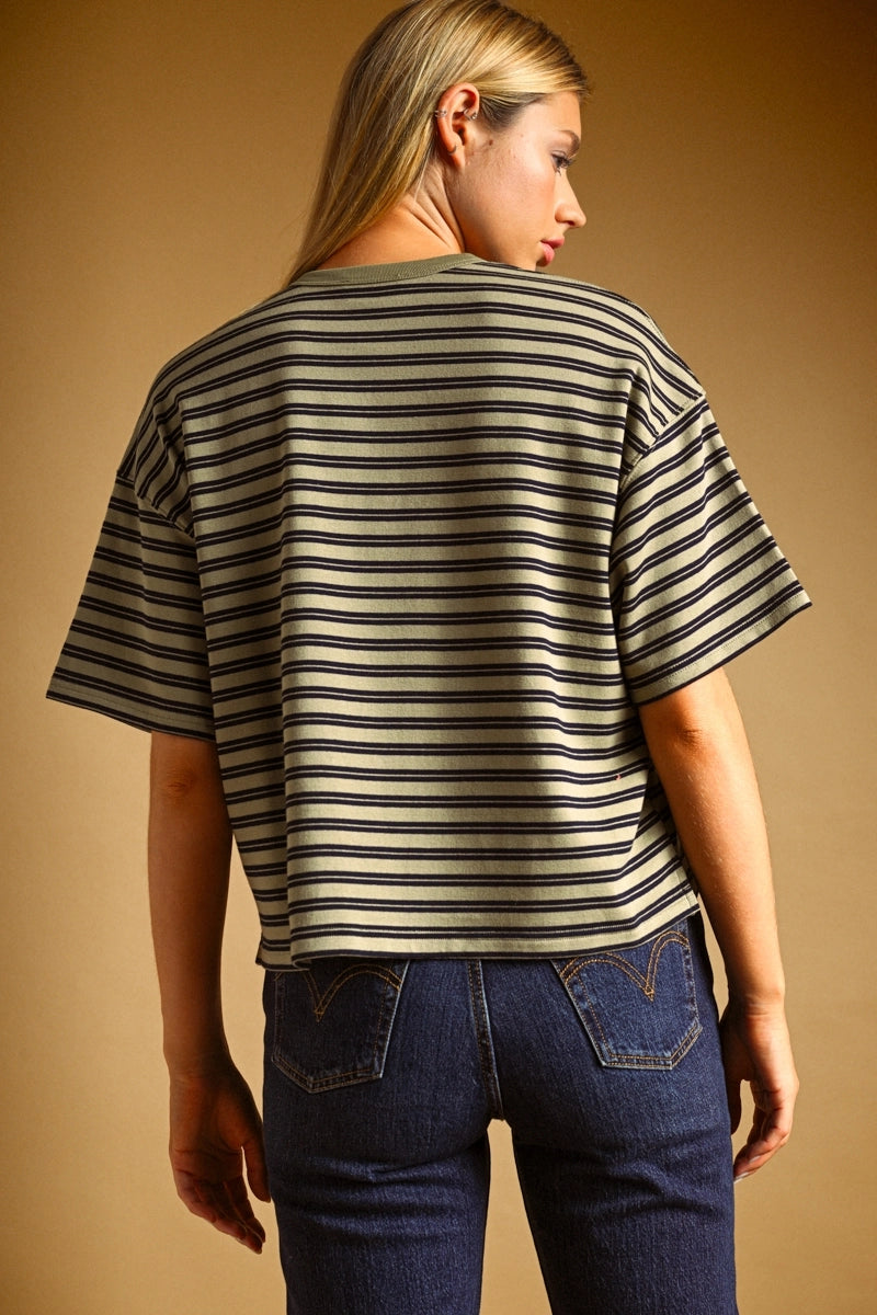 A woman shown from the back wearing a cropped short-sleeve striped t-shirt in olive green and black, paired with dark blue jeans, against a solid warm brown background.