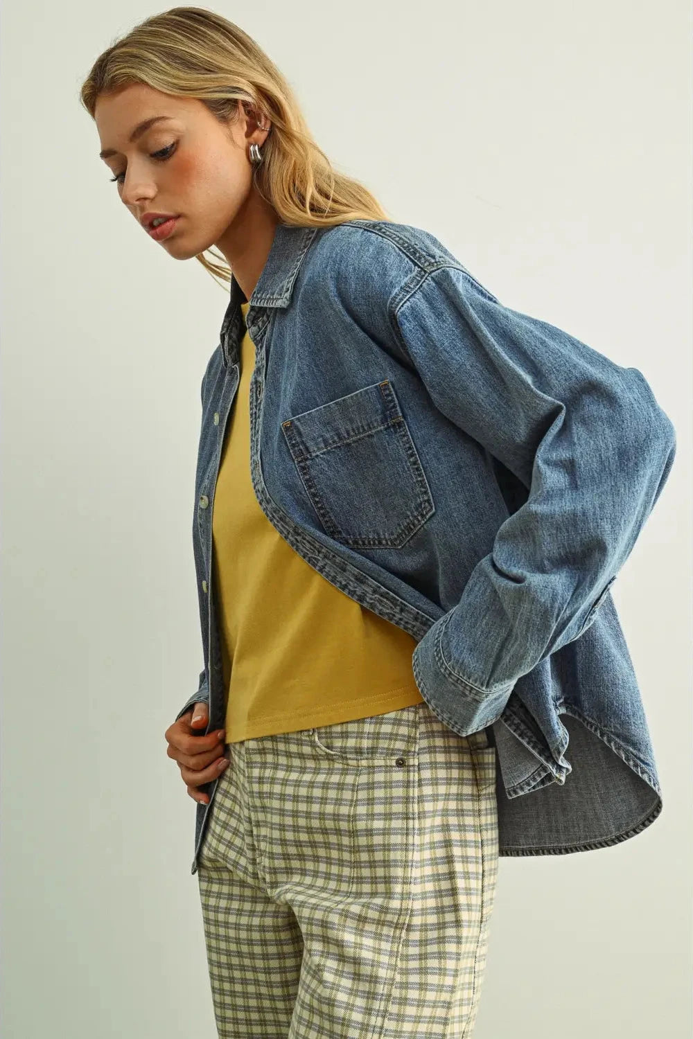 Woman wearing a denim jacket, mustard yellow top, and plaid pants on a light background