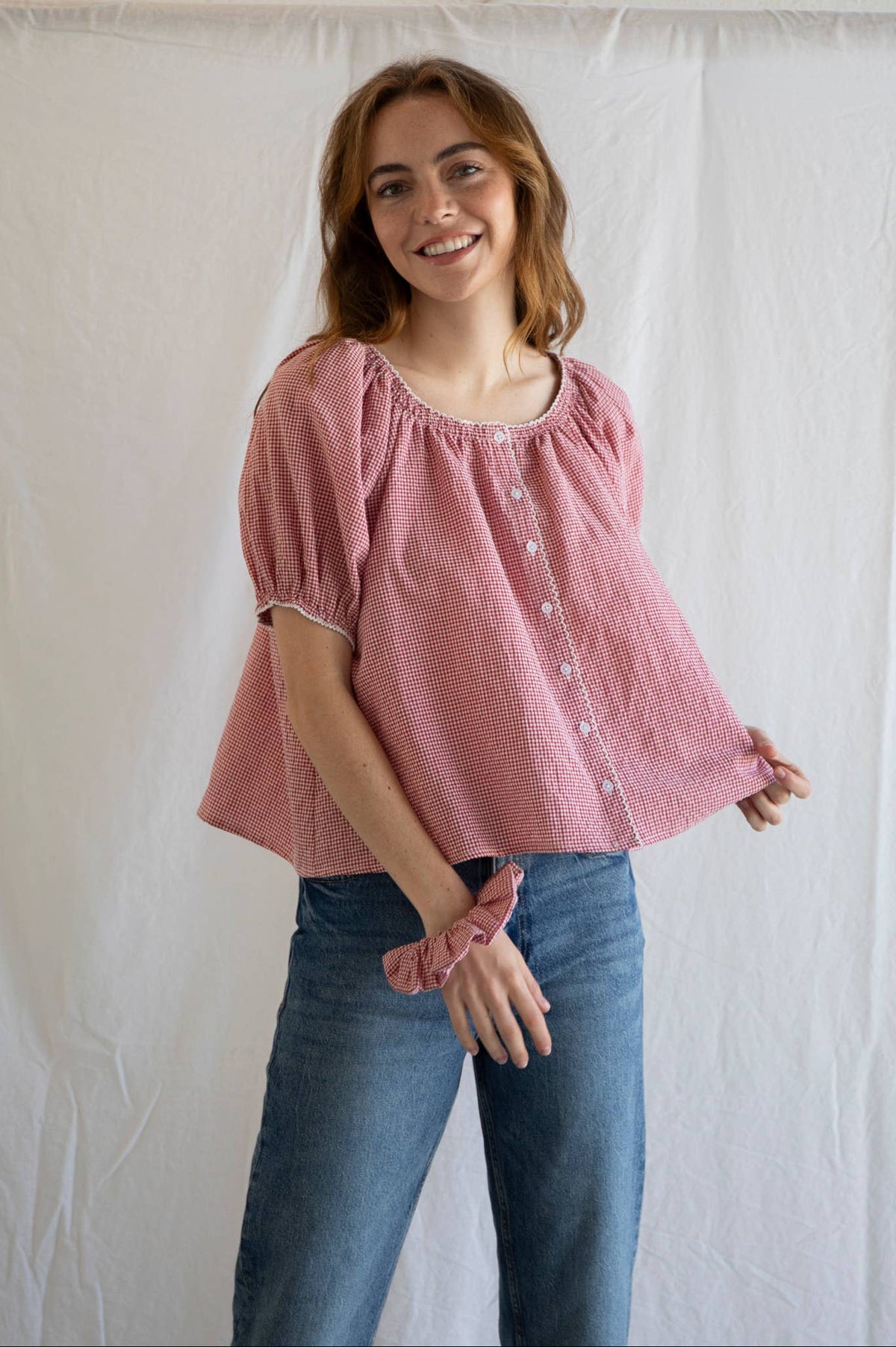 Woman wearing a pink blouse and blue jeans standing against a white curtain.
