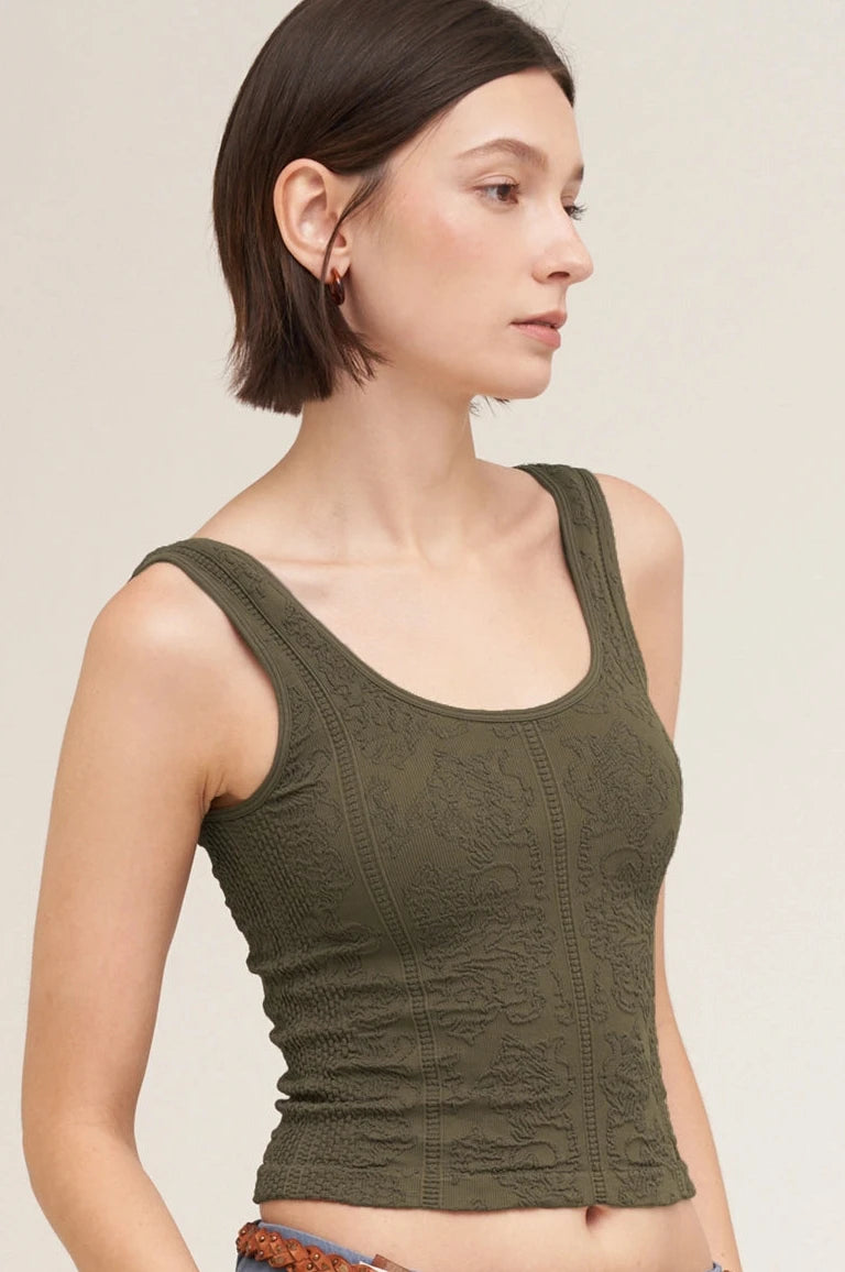 A woman wearing a textured olive green ribbed tank top and blue jeans with a braided brown belt, shown against a plain light beige studio background.