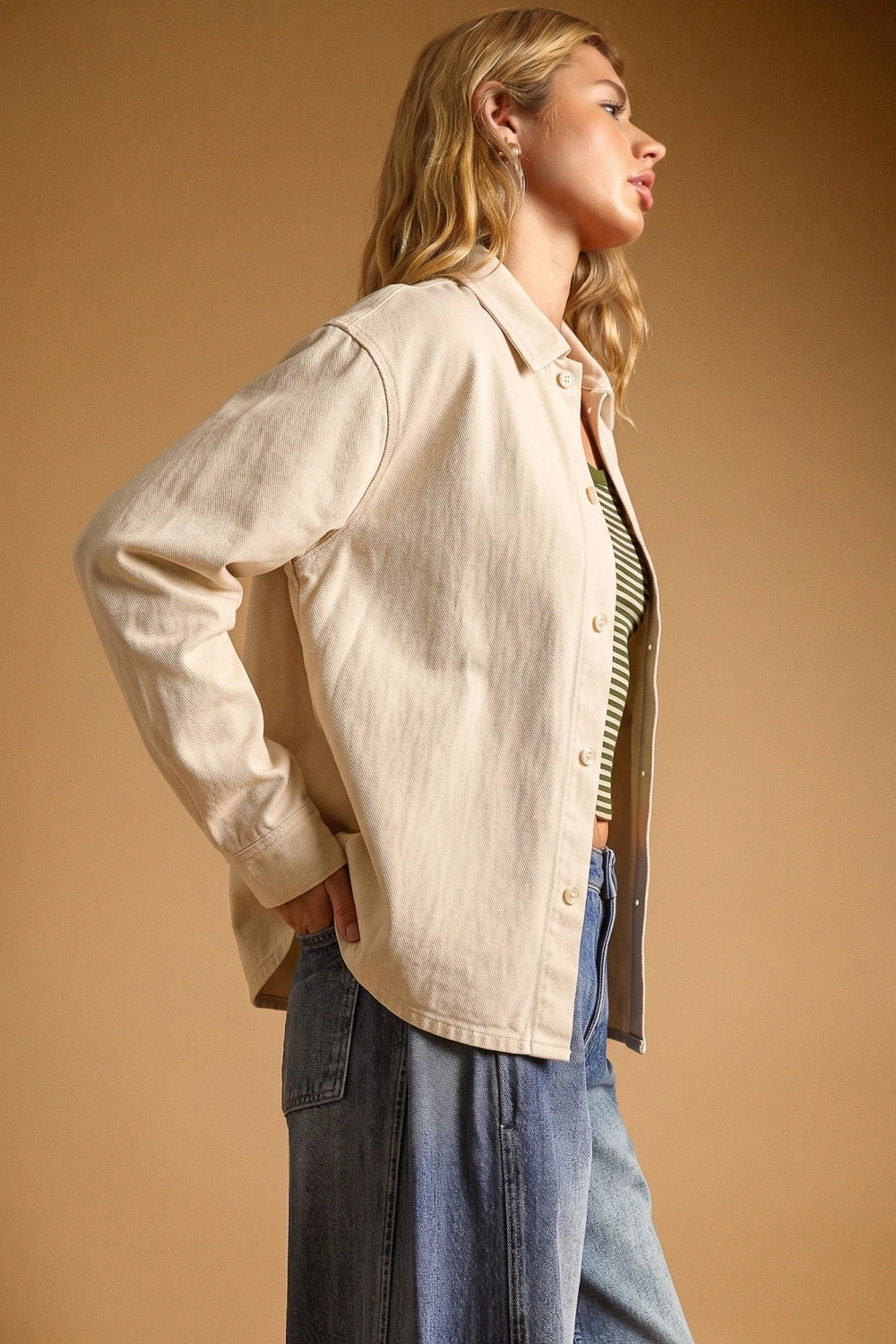 A woman wearing a light beige linen button-up shirt jacket over a green striped top and blue jeans, posed against a solid warm tan background.