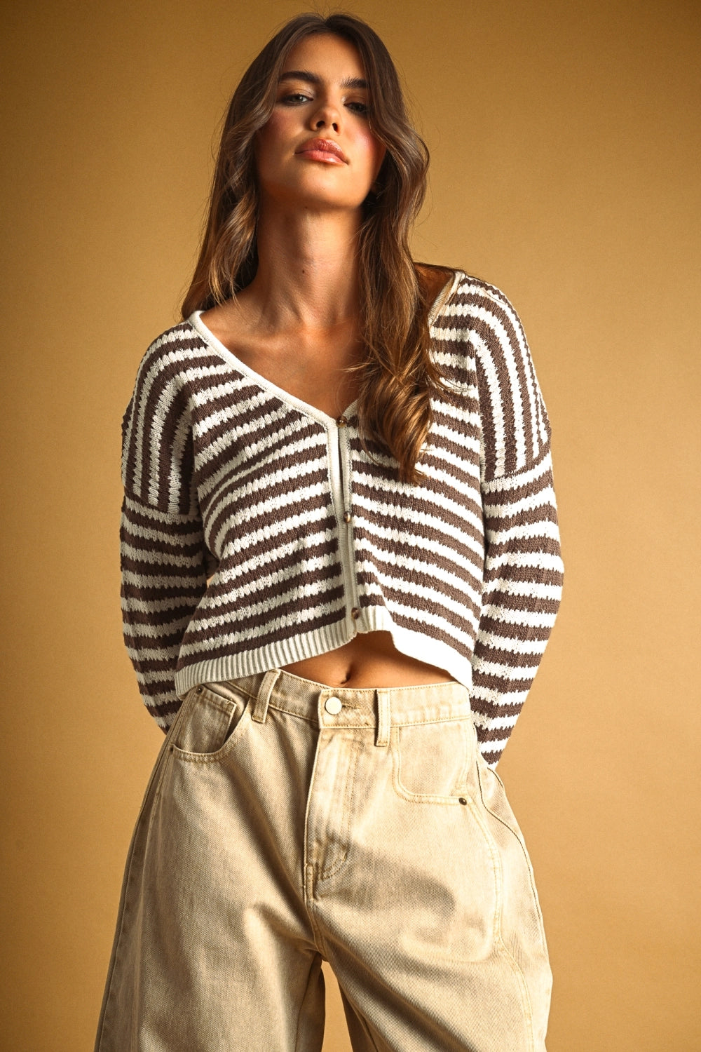 A woman wearing a brown and white striped cropped cardigan sweater and beige wide-leg jeans, posing against a solid warm tan background.