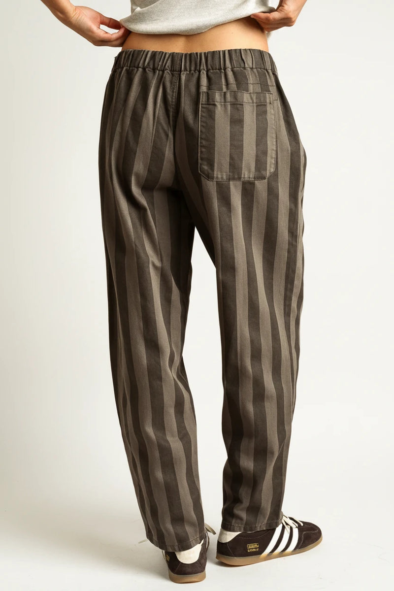 A person shown from the back wearing loose vertical-striped trousers in dark gray and black, paired with black sneakers, against a solid warm tan studio background.