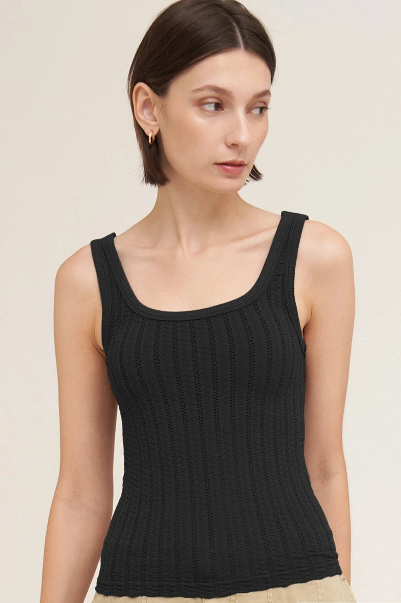 A woman wearing a black ribbed knit scoop-neck tank top, paired with beige pants, against a plain light beige studio background.