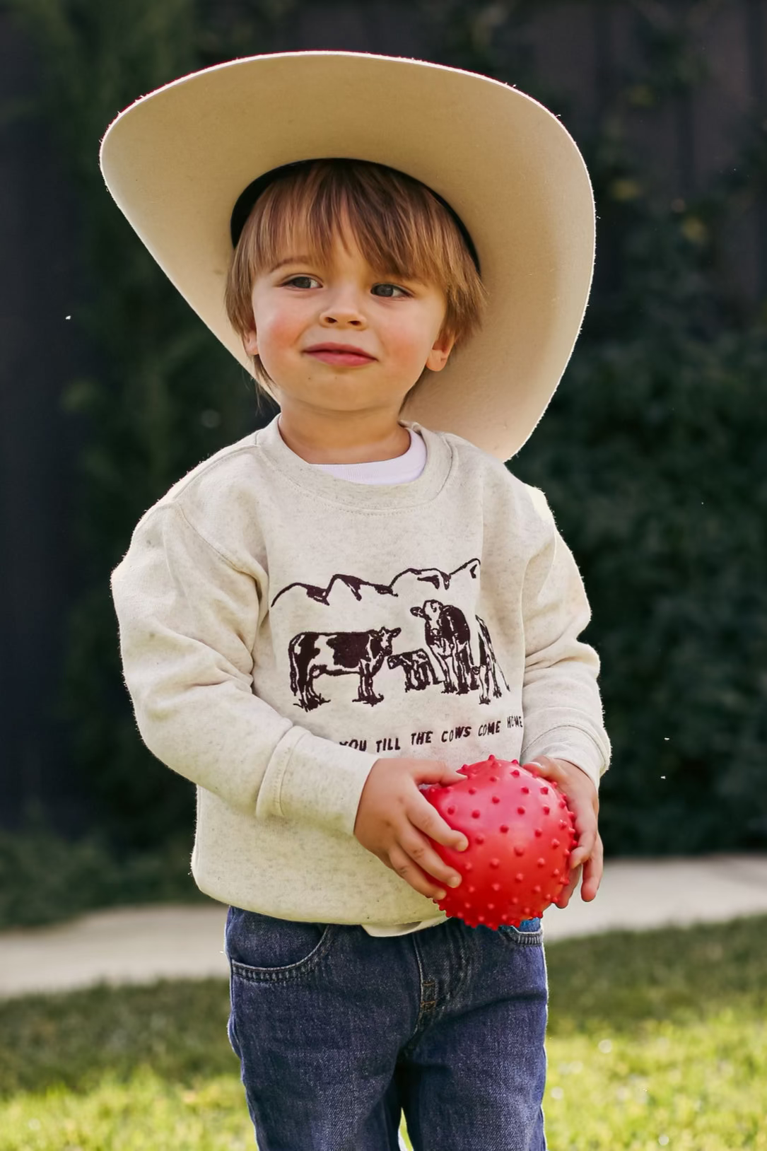 Picture of a toddler boy standing with a sand color crewneck sweatshirt on with a scene of 3 cows and a baby cow embroidered on the front of the sweatshirt with the words Love You Till The Cows Come Home Written underneath.