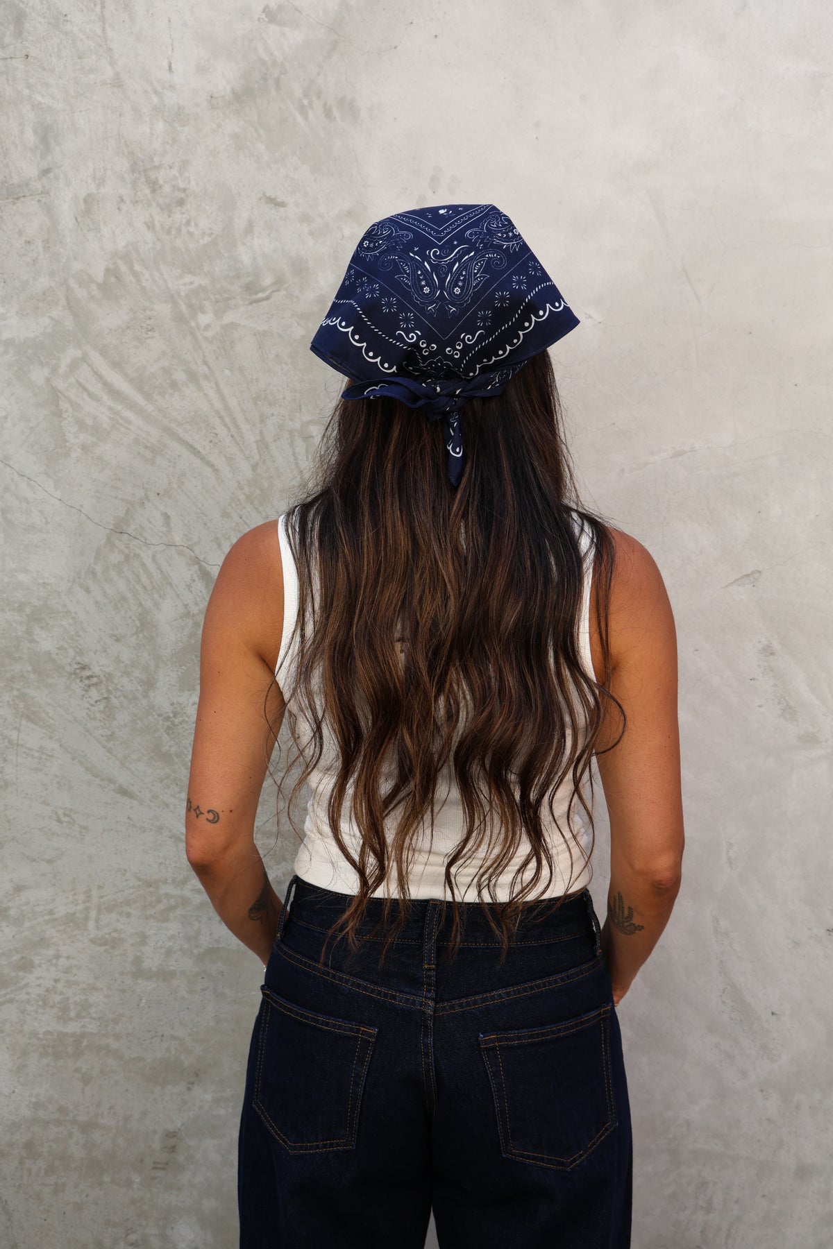 Woman standing wearing a bandana on her head with a white shirt and jeans against a grey wall.