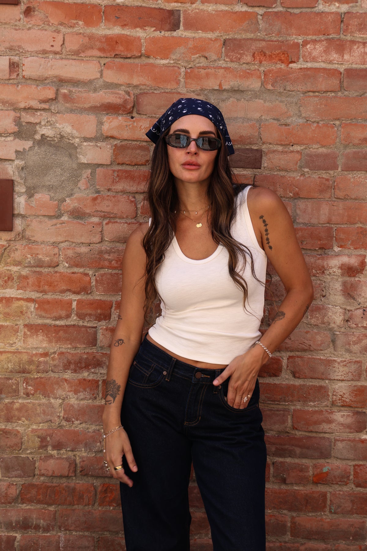 Woman standing wearing a bandana on her head with a white shirt and jeans against a brick wall.