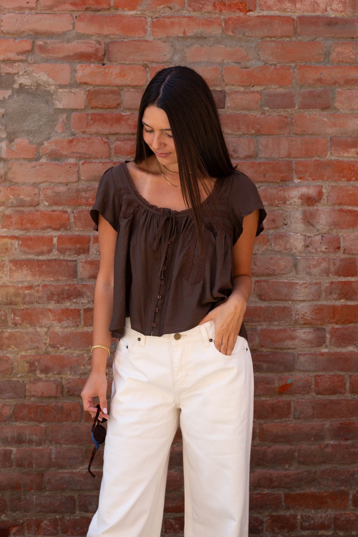 A woman with long dark hair stands against a red brick wall, wearing a dark brown short-sleeve blouse with a gathered neckline and white wide-leg pants.