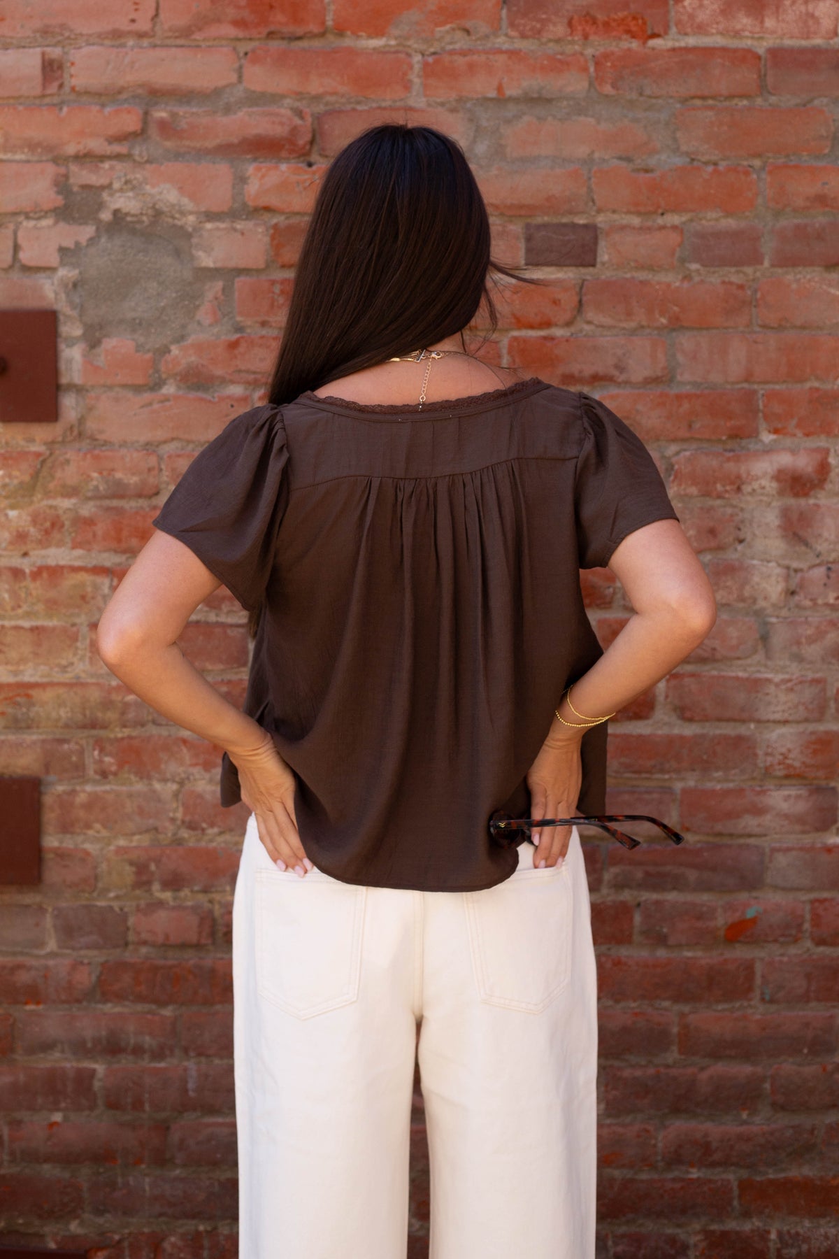 A woman viewed from behind wearing a dark brown short-sleeve blouse with a gathered back neckline and white pants, standing against a red brick wall.