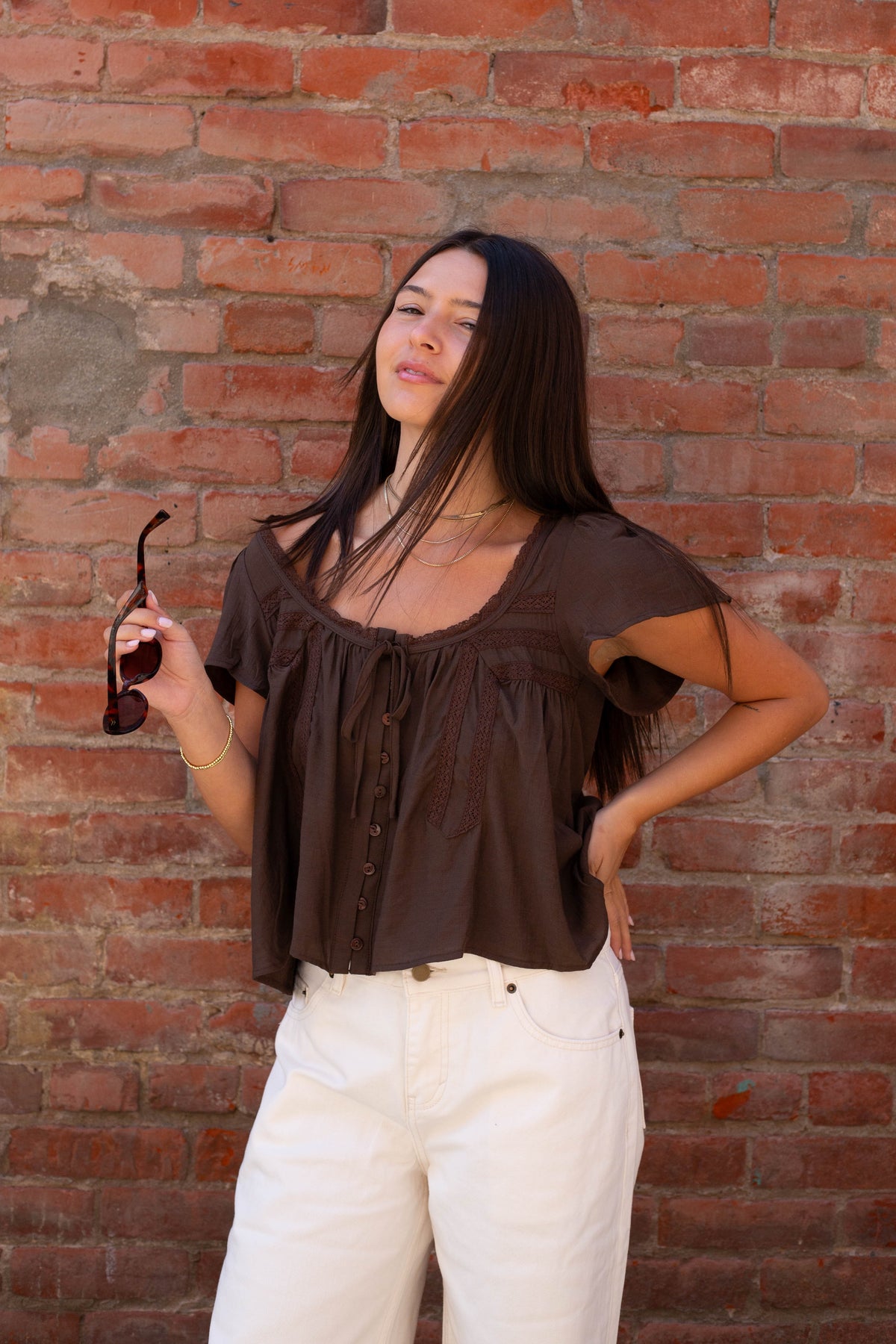 A woman with long dark hair stands against a red brick wall, wearing a dark brown short-sleeve top with a tie detail at the neckline and white pants. She is holding a pair of sunglasses in her right hand.