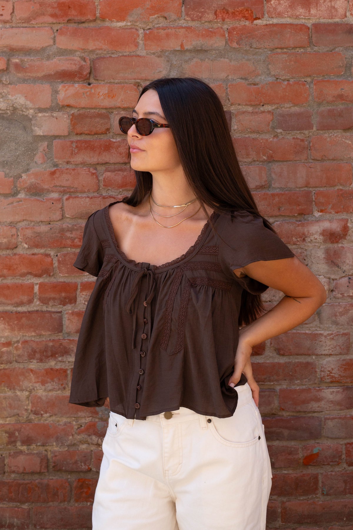 A woman with long dark hair and sunglasses wearing a dark brown ruffled short-sleeve blouse and white pants, standing against a red brick wall.