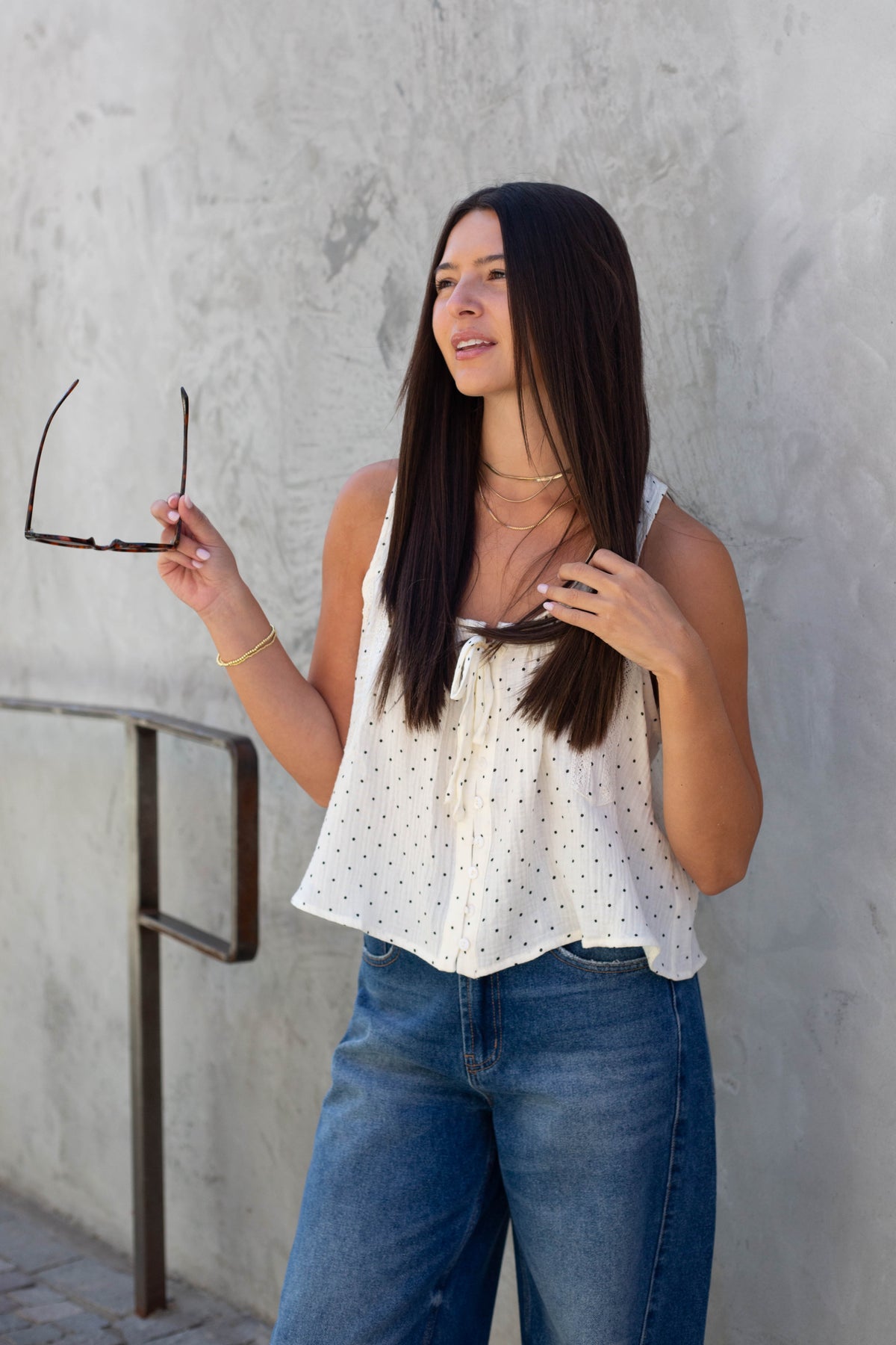 A woman with long dark hair stands against a textured light gray wall, wearing a white sleeveless tie-front top with small black polka dots and blue jeans. She is holding a pair of sunglasses in one hand.