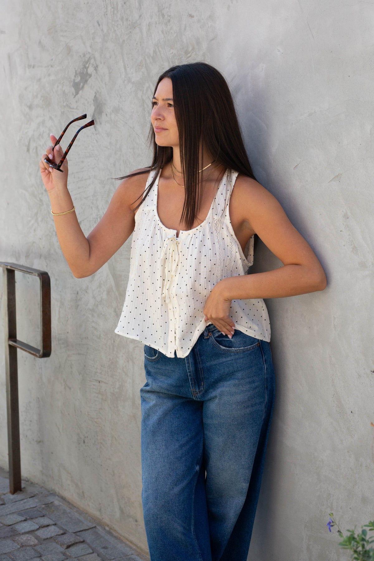 A woman with long dark hair leans against a textured gray concrete wall, wearing a white sleeveless top with tiny black polka dots and blue jeans. She is holding a pair of dark sunglasses in her right hand.