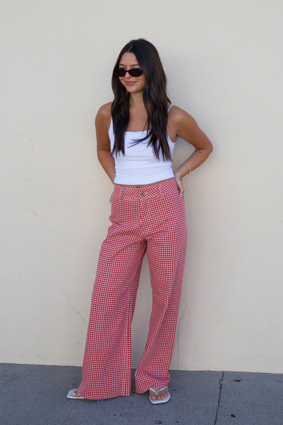 A woman wearing red and white gingham wide-leg pants, a white tank top, and sunglasses, posing against a plain beige wall.